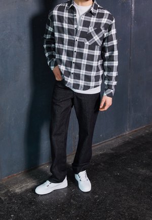 Mand iført sort-hvid ternet skjorte, sorte bukser, hvide sneakers, stående op ad en mørk struktureret væg med den ene hånd i lommen.