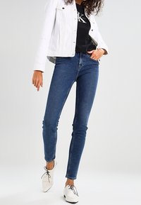 Blå skinny jeans med hög midja, tillsammans med en svart t-shirt och en vit denimjacka. Vita skor med svarta snören.