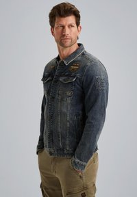 Veste en denim de couleur bleu foncé avec une finition délavée. Présente deux poches poitrine, des boutons en métal et des motifs brodés sur les épaules.