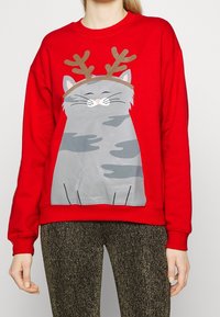 Sudadera roja con un dibujo de un gato de dibujos animados gris con cuernos de ciervo impreso en el frente. La tela es suave con puños y cintura acanalados.