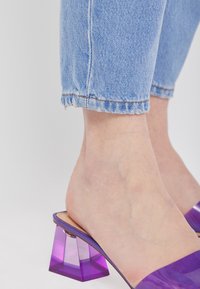 Las sandalias de slide moradas presentan un tacón de bloque de acrílico transparente y una parte superior suave y lisa, combinadas con jeans de mezclilla azul con dobladillo deshilachado.