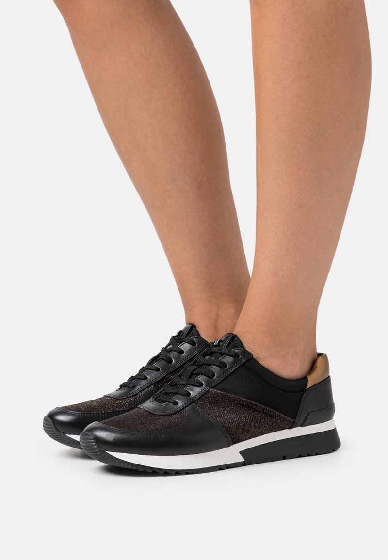 Baskets basses noires avec lacets et semelles blanches portées pieds nus, avec des côtés texturés et un petit détail métallique au talon.