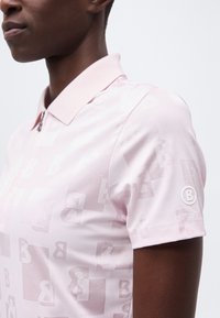 Gros plan d'une personne portant un polo rose clair avec un motif subtil en "B" et un logo rond "B" sur la manche gauche.