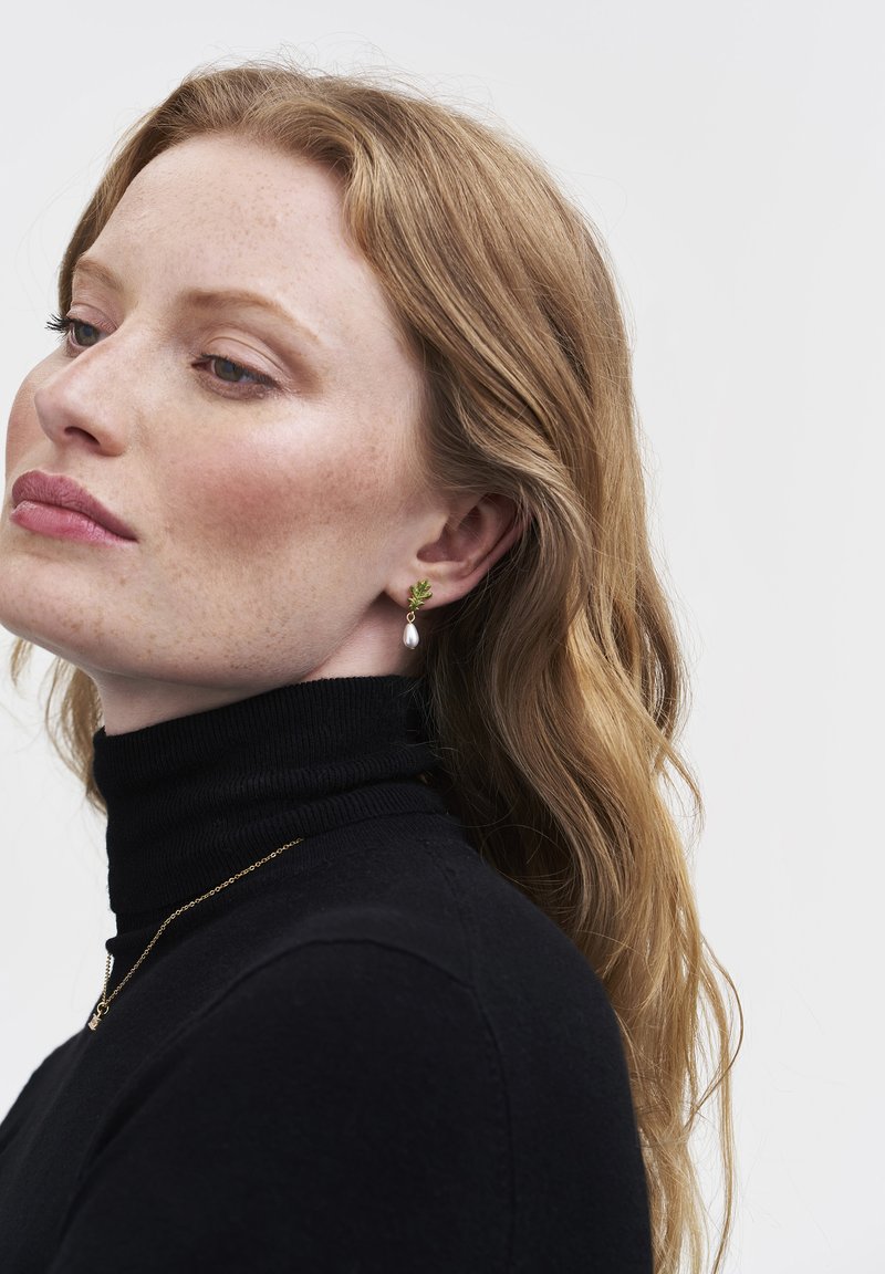 Zelta tonī auskari ar zaļas lapas akcentu un bumbierveida pērles piekariņu, valkāti ar melnu augstu kaklu svārku.