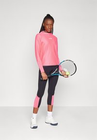 Maglietta sportiva a maniche lunghe rosa con cappuccio, abbinata a leggings neri con dettagli rosa. Tiene in mano una racchetta da tennis con una pallina.