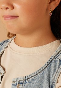 Gouden oorring in een cirkelvorm met een hartvormige roze edelsteen, gezet in een getextureerde, gedetailleerde rand, gedragen door een kind met een spijkerjack en een crèmekleurig shirt.