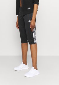 Osoba oblečená v černých adidas sportovních kalhotách ke kolenům s bílými pruhy a bílých sportovních teniskách, stojící proti jednobarevnému pozadí.