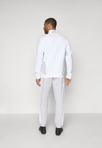 Felpa sportiva bianca con collo alto e polsini a costine, abbinata a pantaloni della tuta grigi chiari con strisce laterali e gamba affusolata.