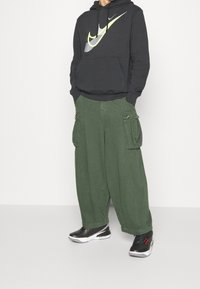 Musta huppari, jossa suuri keltainen ja harmaa Nike-logo, yhdistettynä löysiin vihreisiin cargohousuihin, joissa on sivutaskut. Mustat lenkkarit viimeistelevät asun.