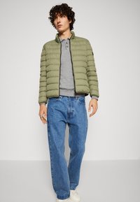 Giacca imbottita verde oliva con colletto alto, cerniera nera, indossata sopra una polo grigia. Abbinata a jeans in denim blu larghi e sneakers bianche.