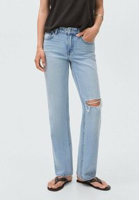 Jeans de mezclilla azul claro con un corte relajado, que presentan un detalle desgastado prominente en la rodilla derecha y un dobladillo desflecado. Combinados con una blusa negra.