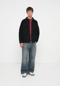 Black denim jacket with contrasting collar, two front pockets, paired with loose-fitting light blue jeans and white sneakers. Solid maroon shirt beneath.
