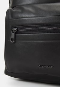 Svart lærryggsekk med en frontlomme med glidelås, glatt tekstur, og "Calvin Klein"-logo preget på siden.