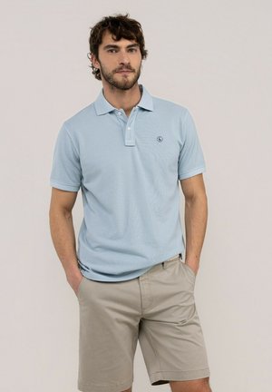 Hombre con cabello corto y rizado y barba, vistiendo una camiseta tipo polo azul claro y pantalones cortos beige, de pie con las manos en los bolsillos.