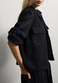 Giacca di jeans nera con vestibilità rilassata, cuciture in vista, due tasche sul petto e maniche arrotolate. Tessuto liscio e forma strutturata.