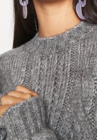 Šedý pletený svetr s texturovanými vzory a vroubkovanými manžetami; má vysoký límec a je doplněn průhlednými náušnicemi.
