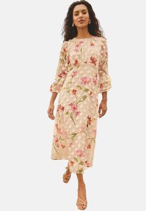 Žena v svetlom béžovom midi šatách s 3D kvetinovou textúrou a ružovo-zelenými kvetinovými potlačami, v kombinácii s telovými sandálmi na remienky.