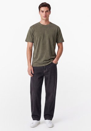 Mann steht mit der Hand in der Tasche, trägt ein olivgrünes T-Shirt mit kurzen Ärmeln, lockere schwarze Jeans und weiße Sneakers vor weißem Hintergrund.