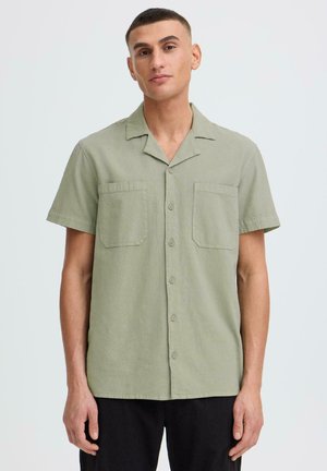 Homme portant une chemise boutonnée à manches courtes de couleur vert olive avec deux poches poitrine et un pantalon noir, debout devant un fond uni.