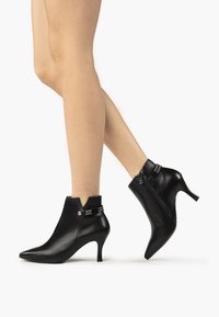 Zwarte leren enkelboots met puntige neuzen en hoge hakken, voorzien van een accent rits en een decoratieve band met versieringen.