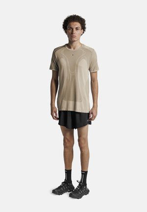 Jonge man staand, gekleed in een beige sportshirt met korte mouwen, zwarte korte broek, zwarte sokken en zwarte hardloopschoenen op een witte achtergrond.