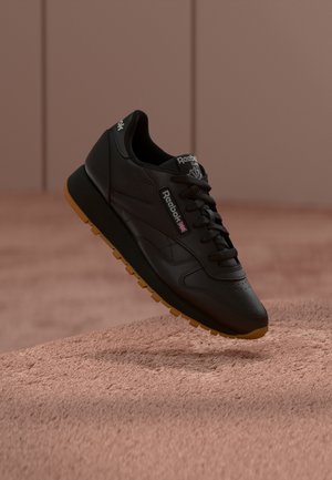 Svart Reebok Classic-sneaker med gummisåle som svever over en teksturert beige overflate mot en myk brun bakgrunn.