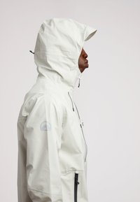 Giacca impermeabile bianca con cappuccio regolabile, chiusura zip sul fronte, tasche laterali e un logo impresso sulla manica sinistra. Tessuto liscio.