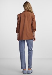 Blazer marron à coupe ajustée, manches longues et col cranté, associé à un jean bleu clair à jambe droite et des chaussures à semelles souples.