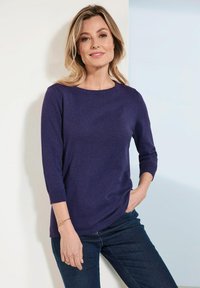 Lila Strickpullover mit rundem Halsausschnitt und 3/4-Ärmeln, kombiniert mit dunklen Jeans, vor einem hellen Hintergrund.