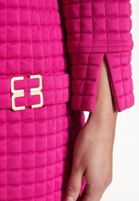 Světlé růžové prošívané látky s texturovaným, polstrovaným vzhledem. Zahrnuje zlatou obdélníkovou sponu a detail manžety na rukávu.