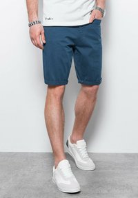 Pantalones cortos de algodón azules con pu�ños enrollados, que cuentan con bolsillos laterales y un cierre frontal. Combinados con zapatillas deportivas blancas de diseño texturizado.