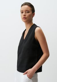 Zwarte mouwloze blouse met een V-hals, gemaakt van een gestructureerde stof, met een ontspannen pasvorm. Gecombineerd met lichtgekleurde broeken.