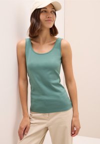 Top aderente verde con scollatura a barca, realizzato in tessuto morbido. Abbinato a pantaloni chiari e un cappellino bianco. Design semplice.
