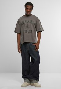 Pelēka pārāk liela T-krekls ar melniem "KARLKANI NY_CUSA" uzrakstiem, saskaņots ar tumši ziliem platām kājām džinsiem un krēmkrāsas kedām.