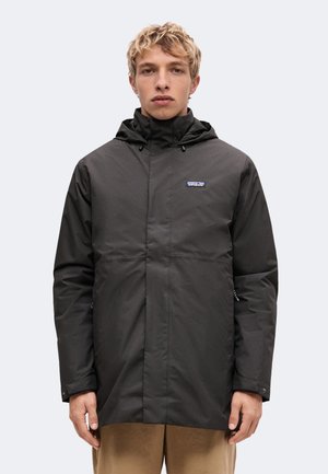 Junger Mann mit kurzen blonden Haaren, der eine schwarze Kapuzenjacke von Patagonia und beige Hose trägt, steht vor einem einfachen hellen Hintergrund.