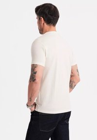 Cremefarvet t-shirt med korte ærmer og rund halsudskæring, lavet af blødt stof. Har synlige tatoveringer på armene og bæres med mørke jeans.