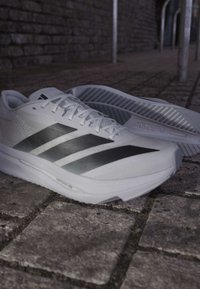 Άσπρα και μαύρα παπούτσια τρεξίματος Adidas πάνω σε πλακόστρωτο, το ένα παπούτσι όρθιο και το άλλο στο πλάι δείχνοντας την σόλα.
