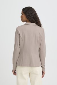 Mujer con cabello largo y rizado, vestida con un blazer beige y pantalones crema, vista desde atrás sobre un fondo blanco liso.