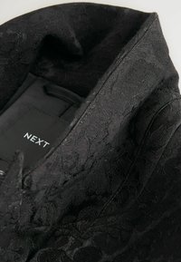 Black textured fabric with floral pattern, featuring a pointed collar and a care label attached inside the garment.