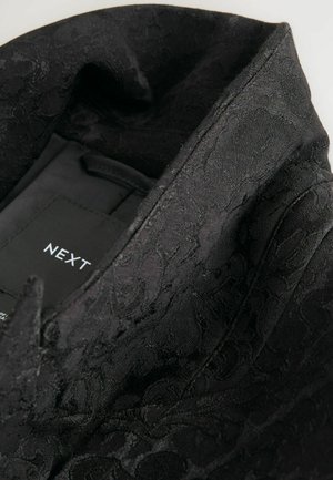 Fekete, mintás anyag virágmintával, hegyes gallérral, belső címkével.