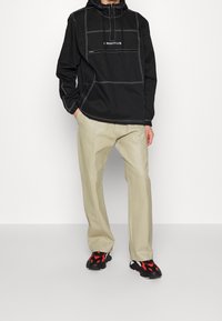 Personne portant une veste à capuche noire avec des coutures blanches, un pantalon beige et des baskets noires avec des accents rouges, debout devant un fond blanc uni.