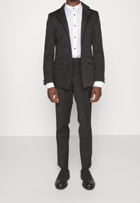Uomo in un completo nero su misura con una camicia a colletto bianco e scarpe eleganti in pelle nera, in piedi davanti a uno sfondo semplice.