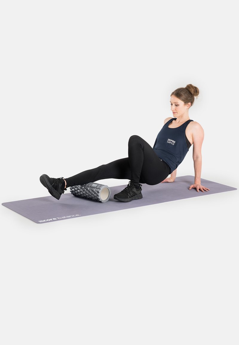 Gray exercise mat with a textured surface, a black foam roller with ridges, and a person with a bun wearing a dark tank top and leggings.
