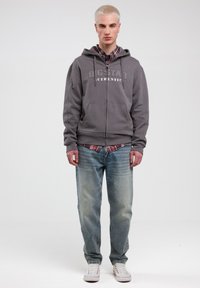 Grå zip-up hættetrøje med frontlommer, prydet med "BIGSTAR AUTHENTIC" logo, lagdelt over en rød ternet skjorte og parret med lyseblå jeans og hvide sneakers.