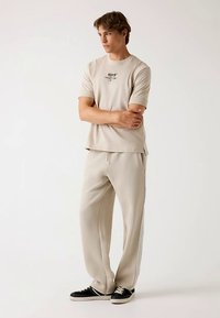 Beżowa koszula z krótkim rękawem z logo, zestawiona z pasującymi beżowymi spodniami z bocznymi paskami. Model nosi czarne tenisówki.