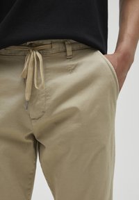 Beiga, lätta chinos med dragsko i midjan, sidofickor och en texturerad yta. Har en subtil sömdetalj på fickan.