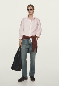 Lichtroze linnen shirt, donkerbruine trui om de taille geknoopt, blauwe spijkerbroek, zwarte tas, bruine suède schoenen, zonnebril. Schone, minimalistische uitstraling.