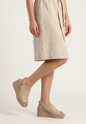 Beine in beigem knielangem Kleid und passenden Keilsandalen mit offenen Zehen und elastischen Rückriemen auf schlicht hellem Hintergrund.