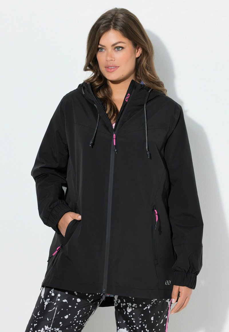 Frau trägt eine schwarze Kapuzenjacke mit rosa Reißverschluss-Details und schwarz-weiß gesprenkelte gemusterte Hose, steht vor einem weißen Hintergrund.