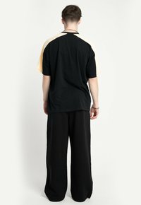 Mężczyzna stojący tyłem, ubrany w czarną i beżową koszulę z krótkim rękawem oraz luźne czarne spodnie na tle jednolitego białego tła.